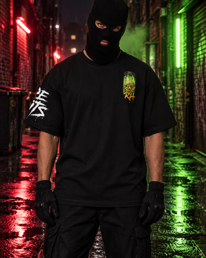 A person wearing a black balaclava, black t-shirt with a green and yellow chest graphic and white sleeve print, and black cargo pants stands in a wet alley at night with red and green neon lights reflecting on the ground.