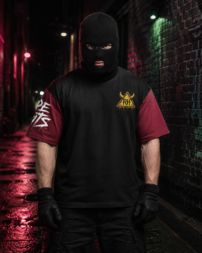 A man wearing a black balaclava mask, black gloves, and a black t-shirt with maroon sleeves stands in a dark alley. The shirt features a yellow triangular logo on the left chest and a white graphic on the right sleeve.