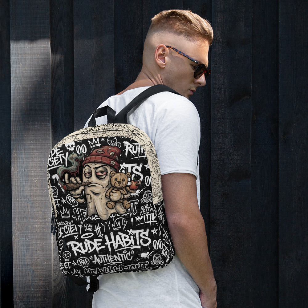 A man wearing a white t-shirt and sunglasses, shown from the back and side, carrying a black backpack with a detailed graphic of a man's face, a teddy bear, and graffiti-style text. The background is a dark wooden plank wall.